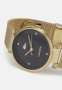 Montre-bracelet en maille dorée avec un cadran noir, présentant des accents argentés, des marqueurs semblables à des diamants et un logo de crocodile à la position 3 heures.