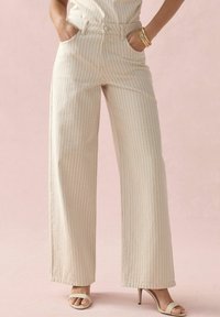 Pantalones de pierna ancha en una tela de beige claro con rayas verticales rojas. Presentan bolsillos frontales y un cierre de botón, combinados con sandalias de tacón bajo claras.