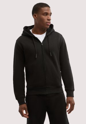 Schwarzer Kapuzenpullover mit Reißverschluss und Kordeln, getragen von einem Mann, der zur Seite schaut, kombiniert mit schwarzen Hosen und einem weißen Hemd darunter.