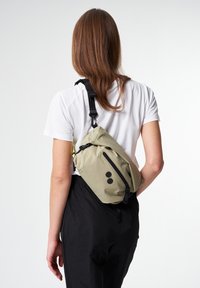 Slingtasche in hellolivgrün, aus strukturiertem Stoff, mit einer Reißverschlusstasche auf der Vorderseite und schwarzen Beschlägen. Verstellbarer Tragegurt für Komfort.