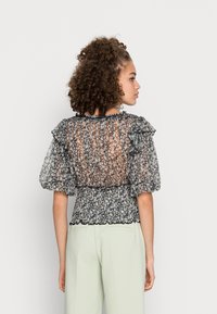 Blusa preta transparente e floral com mangas em babados, cintura franzida e tecido texturizado, combinada com calças verde claro.