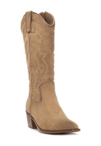 Bota de vaquero suede beige con punta afilada y tacón bajo apilado. Presenta costuras decorativas y un eje ligeramente curvado.
