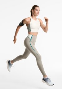 Vrouw die rent in beige en witte sportkleding met teal accenten, draagt lichte grijze sneakers en een zwarte armpouch tegen een witte achtergrond.