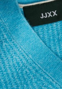 Maglione turchese con un motivo a maglia testurizzata e scollatura a V. L'etichetta presenta testi bianchi su uno sfondo nero.