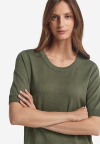Olivgrön stickad tröja med rund halsringning och korta ärmar, med en mjuk textur och ribbade detaljer vid ärmsluten och nederkanten.