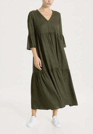 Donna che indossa un vestito midi a balze verde oliva con maniche a campana 3/4 e sneaker bianche, in piedi davanti a uno sfondo chiaro e uniforme.