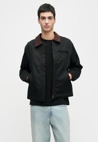 Jaunas vyras vilki juodą striukę su rudomis apykaklėmis, juodą marškinį ir šviesiai mėlynus džinsus, stovintis su rankomis striukės kišenėse.