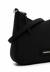 Sac à main noir avec une texture lisse, présentant des coutures apparentes le long des coutures, une fermeture éclair et un accent de logo doré. Bandoulière ajustable.
