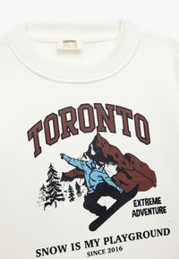 Sudadera blanca con el texto "TORONTO" en marrón oscuro, un gráfico de un snowboarder y el texto "LA NIEVE ES MI PATIO DE JUEGO". Tela suave, escote redondo.