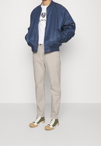 Man som bär en marinblå bomberjacka, vit t-shirt med tryck, beige byxor och gröna och vita högre sneakers, står framför en enkel bakgrund.