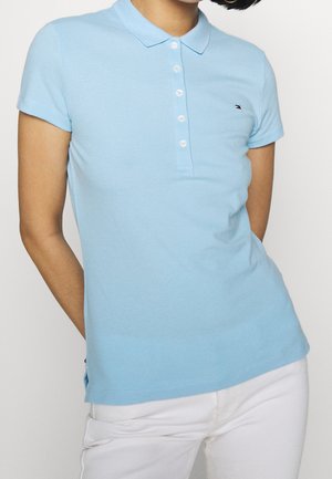 Frau trägt ein hellblaues Poloshirt mit kurzen Ärmeln, weißen Knöpfen und weiße Hose vor einfachem Hintergrund.