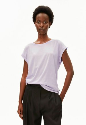 Femme aux cheveux courts et bouclés portant un t-shirt violet clair à manches courtes et un pantalon noir taille haute sur fond blanc.