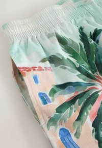 Gros plan d'un short à taille élastique avec un imprimé tropical coloré comprenant des feuilles de palmier vertes et un bâtiment pêche avec des fenêtres bleues.