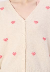 Kremfärgad kofta i mjukt tyg med rosa hjärtan, V-ringning och fem knappar fram. Grov ribbad nederkant.