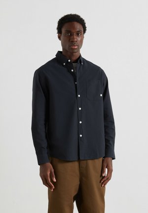 Jonge man in een zwart knoopshirt met borstzak en een broek in bruin, staande tegen een effen lichtgrijze achtergrond.