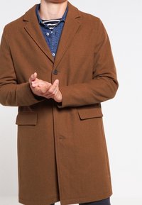 Cappotto in misto lana marrone con rever a ghiera, chiusura a un bottone, due tasche frontali e taglio dritto, indossato sopra una camicia blu.