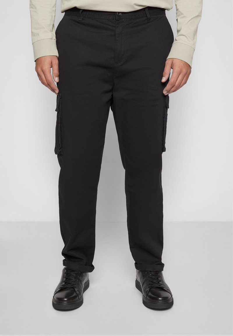 Homme portant un pantalon cargo noir avec des poches latérales et des chaussures noires à lacets, debout devant un fond clair uni.