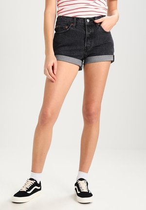 Personne portant un short en jean noir avec revers, des chaussettes blanches aux chevilles, des baskets noires et blanches, et un t-shirt rayé rouge et blanc avec une main dans la poche.