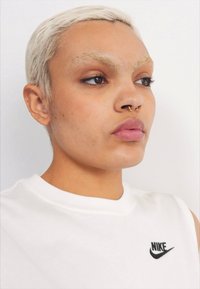 Camiseta sin mangas blanca de Nike con logo negro; cabello corto rubio platino; piercing en el septum visible; piel suave; fondo neutro.