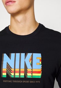 Svart bomull t-shirt med en djärv, flerfärgad "NIKE"-logotyp med 3D-effekt och strukturerad design, inklusive gröna, gula, röda och blå accentfärger.