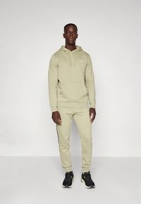 Hellolivgrüner Kapuzenpullover mit Fronttasche und Kordelzug; passende, zulaufende Hose mit seitlichen Streifen; kombiniert mit schwarzen Sportschuhen.