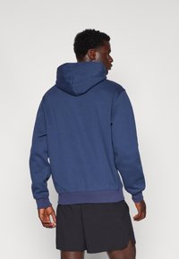 Felpa blu navy con cappuccio regolabile, polsini e orlo a coste, realizzata in materiale morbido. Indossata con pantaloni corti neri per un look informale.