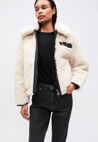 Femeie îmbrăcată cu o jachetă sherpa de culoare crem, cu detalii negre la fermoar, un tricou negru și blugi negri decolorati cu talie înaltă, stând cu o mână în buzunar.