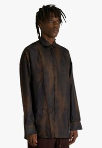 Camicia a maniche lunghe color marrone scuro con sottili righe verticali, chiusura frontale con bottoni e una texture morbida; presenta un colletto classico e una vestibilità ampia.