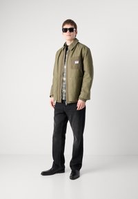 Olive green jacket with a chest pocket, layered over a patterned shirt. Dark jeans and black shoes complete the outfit. Person wearing sunglasses.