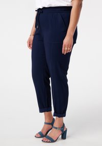 Pantalons navy avec une taille élastique, deux poches latérales et des ourlets retournés. Associés à des sandales à talons bleues avec des brides autour des chevilles.