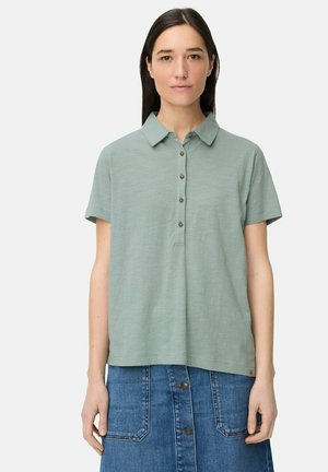 Ung kvinde iført en kortærmet salviegrøn knappet polotrøje og en højtkwaistet blå denimnederdel med store forlommer.