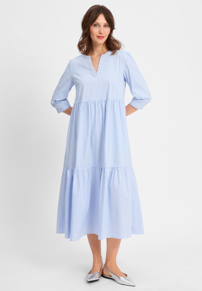 Hellblau gestreiftes Kleid mit V-Ausschnitt, dreiviertelärmel und einem ausgestellten Rock. Getragen mit silbernen Ballerinas. Glatte Stoffoberfläche.