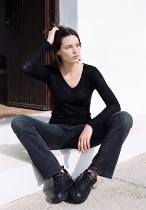 Une femme vêtue d’un haut noir, d’un jean foncé et de bottes noires est assise sur des marches blanches, se tient les cheveux et regarde sur le côté.