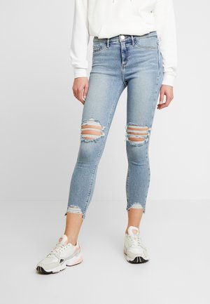 Osoba oblečená v světle modrých roztrhaných úzkých džínách, bílých teniskách s růžovými akcenty a bílém dlouhém tričku stojící na šedé podlaze.