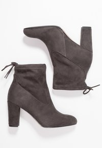 Botines de ante gris con punta afilada, tacón cuadrado de 10 cm y detalle de cordón ajustable en el tobillo, que presentan acentos cosidos.
