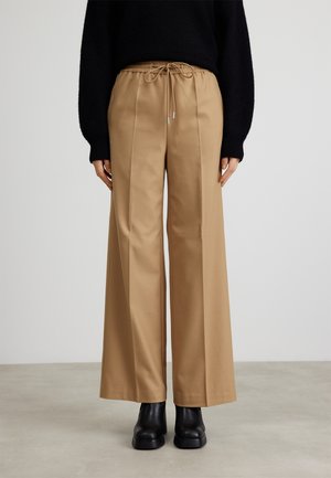 Pantalon beige à jambes larges avec taille élastique à cordon, porté avec un pull noir et des bottines noires sur un fond neutre.