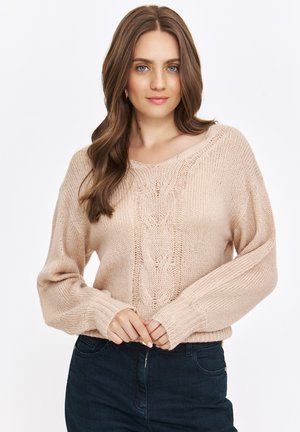 Jasnoróżowy sweter dzianinowy z wzorem warkoczowym, szerokimi rękawami i ściągaczem na dole. Modelka nosi ciemne dżinsy, podkreślając teksturę swetra.