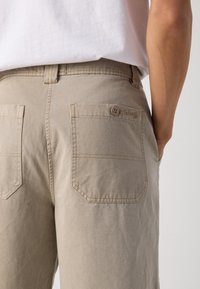 Beige cotton pants with two back pockets, featuring a button closure and stitched detailing. Logo visible on the back pocket.