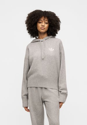 Mujer con cabello rizado que lleva una sudadera con capucha gris clara y pantalones deportivos a juego, de pie frente a un fondo blanco liso.