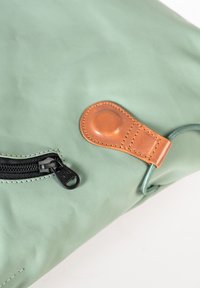 Borsa in tessuto impermeabile verde con una cerniera nera, un accentuazione in pelle beige e un cordino arrotolato. Texture liscia e design minimalista.