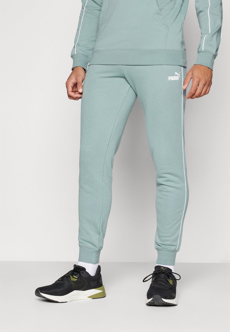 Pantalons de jogging vert clair avec des rayures blanches sur les côtés et un logo PUMA, fabriqués en tissu doux. Associés à des chaussures de sport noires et des chaussettes blanches.
