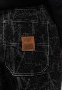 Čierna látková vrecko s bielym prešívaním a abstraktným šedým vzorom, s malou hnedou koženou náplasťou s reliéfnym dizajnom.