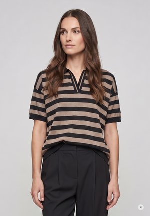 Woman with long wavy brown hair wearing a black and beige horizontal striped short sleeve top and black pants, standing against a gray background.
