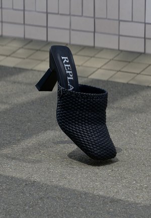 Mule noire tissée à talon avec bout carré et talon bloc, portant l'inscription "REPLA" à l'intérieur, posée sur une surface extérieure en carrelage et bitume.