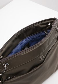 Šedá nylonová taška s modrým vnitřkem, zapínáním na zip a kruhovými kovovými doplňky. Má vnější kapsu a detail loga.