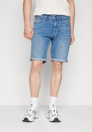 Man draagt lichtblauwe opgerolde spijkershorts, witte sneakers, witte crew sokken en een wit overhemd tegen een effen achtergrond.