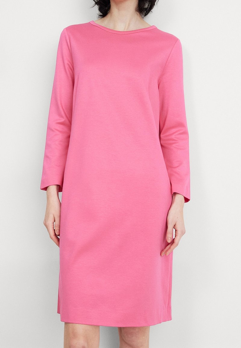 Femme portant une robe rose unie, à manches longues, longueur genou, avec un col bateau, sur fond blanc.