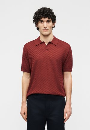 Νέος με κατσαρά μαλλιά, φορώντας μια υφασμάτινη κοντομάνικη πόλο μπλούζα σκούρου κόκκινου χρώματος και μαύρο παντελόνι, στέκεται μπροστά σε ένα απλό λευκό φόντο.