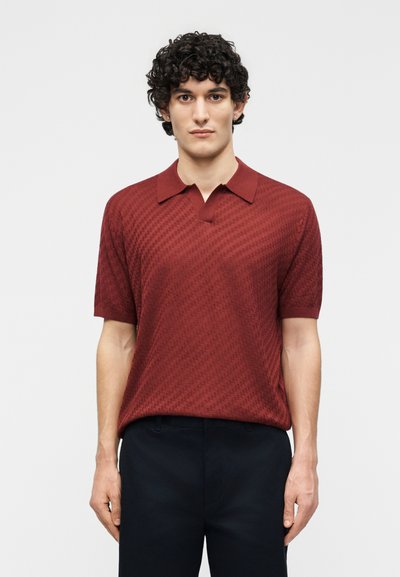 Jeune homme aux cheveux bouclés portant un polo à manches courtes rouge foncé texturé et un pantalon noir, debout devant un fond blanc uni.