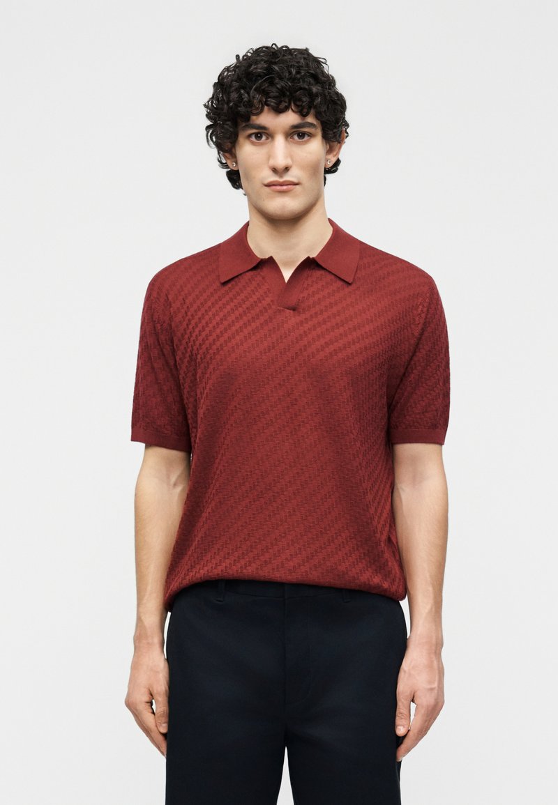 Νέος με κατσαρά μαλλιά, φορώντας μια υφασμάτινη κοντομάνικη πόλο μπλούζα σκούρου κόκκινου χρώματος και μαύρο παντελόνι, στέκεται μπροστά σε ένα απλό λευκό φόντο.